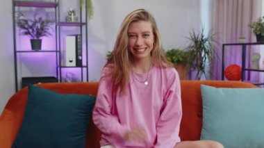 Portrait of caucasian adult girl smiling friendly at camera and waving hands gesturing hello, hi, greeting or goodbye, welcoming with hospitable expression. Young woman indoor at home in room on couch