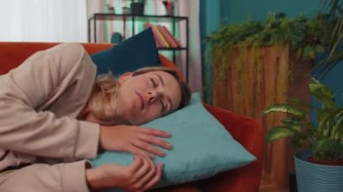 Tired caucasian adult girl lying down in bed taking a rest at home. Carefree young woman napping, falling asleep on comfortable orange sofa with pillows. Closed her eyes enjoy daytime nap alone
