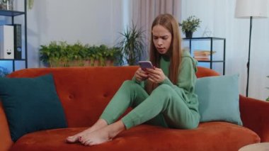 Portrait of caucasian girl use smartphone typing browsing, loses becoming surprised sudden lottery results, bad news, fortune loss, fail. Young child kid sitting on orange couch at home living room