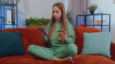 Lovely girl using credit bank card and smartphone while transferring money, purchases online shopping, order food delivery at modern home apartment indoors. Young child kid in living room sit on couch