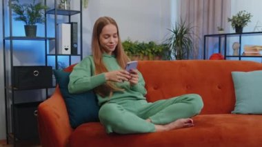 Portrait of girl sitting on sofa uses mobile phone smile at modern home apartment. Young child kid texting share messages content on smartphone social media applications online, watching relax movie