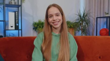 Close-up portrait of happy smiling teen girl in hoodie, looking at camera, celebrate good news. Young child kid indoor isolated at home in living room sitting on orange couch. Female nature beauty