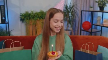 Happy teen girl wears festive birthday hat hold cupcake makes wish joyful congratulating blowing burning candle on cake. Young child kid celebrating anniversary party at home room apartment on couch