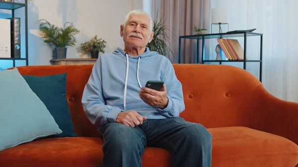 Senior old grandfather sitting on sofa uses mobile phone smile at modern home apartment. Elderly man texting share messages content on smartphone social media applications online, watching relax movie