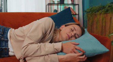 Tired caucasian adult girl lying down in bed taking a rest at home. Carefree young woman napping, falling asleep on comfortable orange sofa with pillows. Closed her eyes enjoy daytime nap alone
