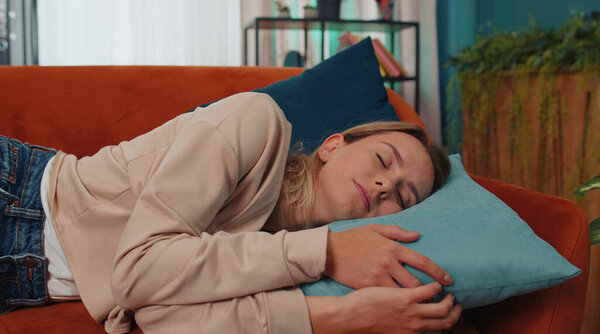 Tired caucasian adult girl lying down in bed taking a rest at home. Carefree young woman napping, falling asleep on comfortable orange sofa with pillows. Closed her eyes enjoy daytime nap alone