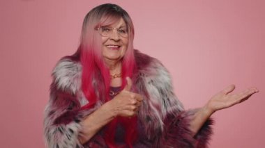 Sevimli yaşlı kadın başparmağını kaldırıp boş yeri işaret ediyor. Ticari metinler için reklam alanı, ürün tanıtımı için fotokopi alanı. Yaşlı büyük anne. Pembe arka planda kapalı stüdyo çekimleri