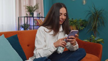 Planning family budget. Smiling caucasian girl counting money cash, use smartphone calculate domestic bills at home. Joyful young woman satisfied of income and saves money for planned vacation, gifts