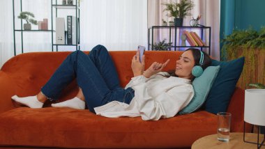 Young woman in wireless headphones relaxing lying on sofa at home choosing listening favorite energetic disco rock n roll dancing music in smartphone. People weekend activities. Happy overjoyed girl