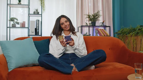 Modern ev apartmanında oturan alışveriş meraklısı genç kadın çevrimiçi yemek siparişi veriyor, kredi kartı ile ödeme yapıyor. İnternette indirimli satış. Kanepede yetişkin bir kız.