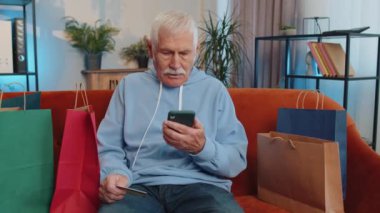 Alışveriş meraklısı, alışveriş poşetleriyle evde oturmuş online sipariş veriyor, kredi kartı ile ödeme yapıyor. Büyükbabam evdeki kanepede yatıyor. İnternette indirimli satış