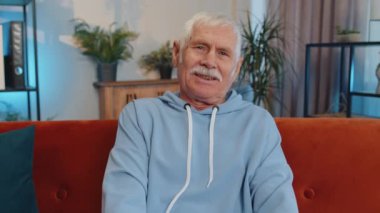 İyi haberi kutlamak için kameraya bakan mutlu, beyaz, yaşlı bir adamın yakın plan portresi. Büyükbabam evde, oturma odasında turuncu koltukta izole halde oturuyor. Erkek doğası güzelliği