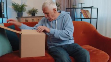 Happy senior grandfather unpacking delivery parcel sitting at home. Smiling satisfied elderly man shopper, online shop customer opening cardboard box receiving purchase gift by fast postal shipping