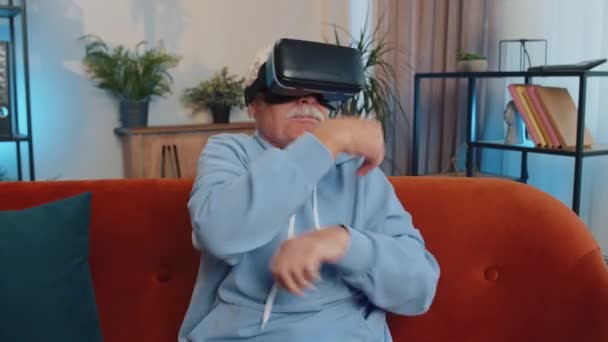 Vrアプリのヘッドセットヘルメットを使用して仮想現実未来的な技術を使用して高齢者の祖父は 現代の家庭のアパートで映画の映画を見て シミュレーション3Dビデオゲームをプレイする 男でゴーグルソファに座って — ストック動画