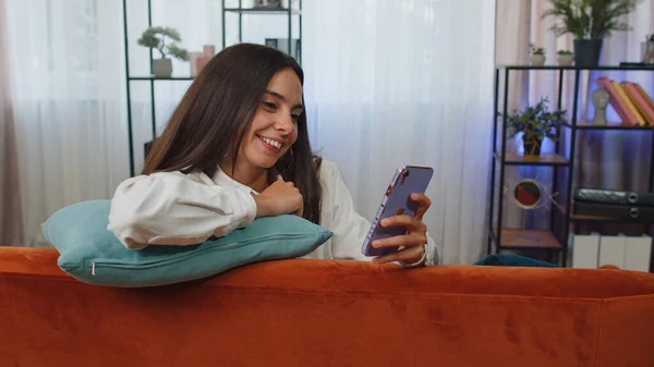 Portrait of adult girl sitting on sofa uses mobile phone smile at modern home apartment. Young woman texting share messages content on smartphone social media applications online, watching relax movie