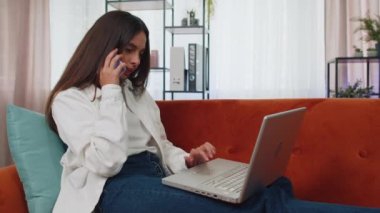 Dizüstü bilgisayar kullanan, cep telefonuyla konuşan, koltukta oturan ev ofisinden çalışan genç bir kadın. Kız yeniden kazanma, internette gezinme, online alışveriş, bilgisayardan yemek siparişi verme