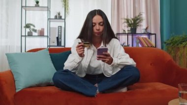 Modern ev apartmanında oturan alışveriş meraklısı genç kadın çevrimiçi yemek siparişi veriyor, kredi kartı ile ödeme yapıyor. İnternette indirimli satış. Kanepede yetişkin bir kız.