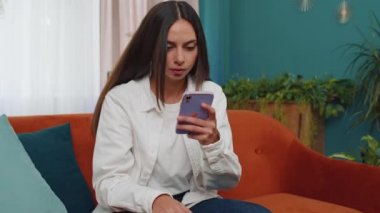 Portrait of caucasian adult girl use smartphone typing browsing, loses becoming surprised sudden lottery results, bad news, fortune loss, fail. Young woman sitting on orange couch at home living room