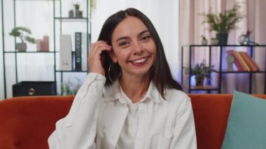 Close-up portrait of happy smiling caucasian girl in shirt, looking at camera, celebrate good news. Young woman indoor isolated at home in living room sitting on orange couch. Female nature beauty