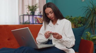 Girl using credit bank card and laptop computer while transferring money, purchases online shopping, order food delivery at home. Young woman sitting on couch holding dollar cash at home living room