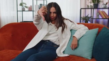 Dislike. Upset girl showing thumbs down sign gesture, expressing discontent, disapproval, dissatisfied bad work at modern home apartment indoors. Displeased young woman in living room sitting on sofa