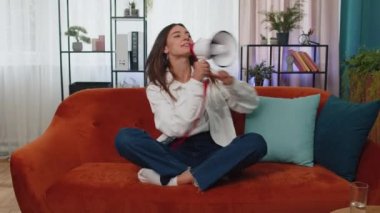 Portrait of adult girl talking with megaphone, proclaiming news, loudly announcing advertisement, warning using loudspeaker to shout speech. Young woman at modern home apartment living room on couch