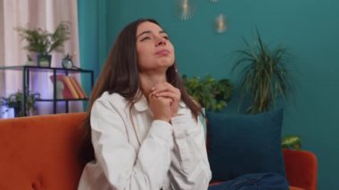 Please, God help. Girl praying sincerely with folded arms, looking upward and making wish, asking with hopeful imploring expression, begging apology. Young woman sitting on couch at home living room