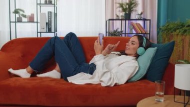 Young woman in wireless headphones relaxing lying on sofa at home choosing listening favorite energetic disco rock n roll dancing music in smartphone. People weekend activities. Happy overjoyed girl