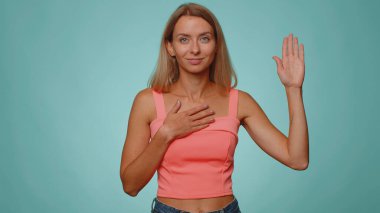 Dürüst olacağıma yemin ederim. Samimi sorumluluk sahibi bir kadın yemin etmek için elini kaldırıyor, dürüst olacağına ve doğruyu söyleyeceğine söz veriyor, elini göğsüne koyuyor. Genç yetişkin bir kız, mavi stüdyo arka planında tek başına.