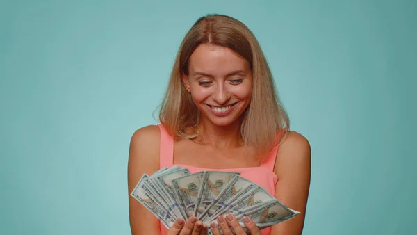 Rich pleased boss blonde woman waving money dollar cash banknotes bills like a fan, success business career, lottery winner, big income, wealth. Young girl isolated alone on blue studio background