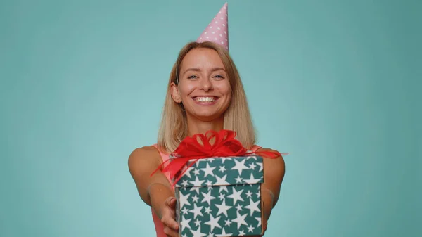 Positive smiling blonde woman presenting birthday gift box stretches out hands, offer wrapped present career bonus, celebrating party. Young adult girl isolated alone on blue studio wall background