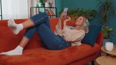 Young woman in wireless headphones relaxing lying on sofa at home choosing listening favorite energetic disco rock n roll dancing music in smartphone. People weekend activities. Happy overjoyed girl