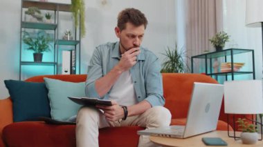 Man accountant working with financial graph documents, calculate taxes for charts and graph reports of home income on couch. Young businessman working on laptop computer, planning expenses budget