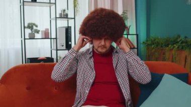 Happy relaxed overjoyed young guy in wireless earphones dancing on couch at home choosing listening favorite energetic disco rock n roll music in smartphone entertaining. People weekend activities