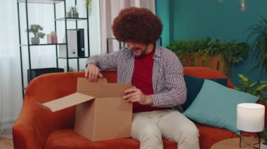 Happy surprised winner adult man unpacking delivery parcel at home. Smiling satisfied young guy shopper, online shop customer opening cardboard box receiving purchase gift ball by fast postal shipping
