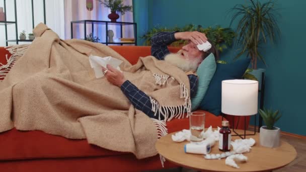 生病的老爷爷 感冒或过敏 躺在家里的沙发上 头戴毛巾的年老爷爷病倒了 打喷嚏把湿巾吹进餐巾里 Coronavirus Quarantine Pandemic — 图库视频影像