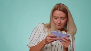 Endişeli, tişörtlü, komik bağımlı kadın cep telefonuyla yarış oyunları oynuyor. Mavi stüdyo arka planında izole edilmiş sürücü simülatörlü akıllı telefon aygıtı uygulaması kullanan genç bir kız