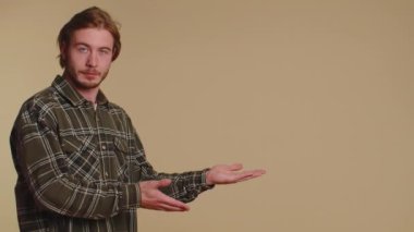 Tişörtlü yakışıklı, iyi niyetli ve boş bir yeri işaret eden ticari metinler için reklam alanı, ürün tanıtımı için fotokopi alanı. Genç bir yetişkin. Bej duvar arka planında çekilen kapalı stüdyo.