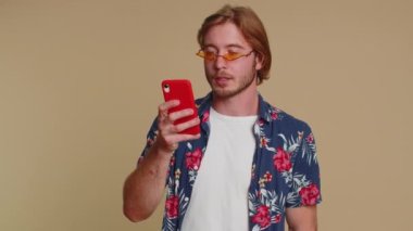 Akıllı telefon görünümlü mutlu turist samimi bir sevinç gösterisi kazanır, para dolar banknotları alır, başarı piyangosunda şans getirir. Genç sakallı çocuk bej renkli stüdyoda tek başına izole edilmiş.