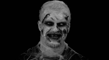 Cadılar Bayramı zombisi makyajlı korkunç bir adam. Korkutmaya çalışıyor, dil gösteriyor. Kasılmalar. Stüdyonun siyah arka planında izole edilmiş uğursuz ölümsüz adam. Voodoo ayinleri. Moda vücut sanatı