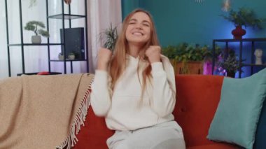 Happy caucasian girl shouting, celebrating success, winning, goal achievement, good news, raising fists in gesture I did it, lottery luck at home apartment. Young woman indoors in living room on couch