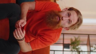 Mutlu kızıl adam bağırıyor, başarıyı kutluyor, kazanıyor, gol başarısı, iyi haber, iyi haber, el hareketleriyle yumruk kaldırıyorum. Odadaki genç adam kanepede. Dikey görünüm