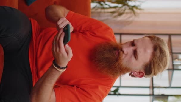 悲しいひげを生やした男は スマートフォンのタイピングブラウジングを使用して 突然の宝くじの結果 悪いニュース 財産の損失 期限に驚いて失う オレンジのソファの上の部屋にいる若い男 縦画面表示 — ストック動画