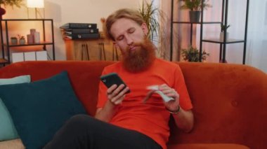 Aile bütçesi planlamak. Sakallı kızıl saçlı adam, evde faturaları hesaplamak için akıllı telefon kullanıyor. Gelirden memnun olan neşeli genç adam planlı tatil ve hediyeler için para biriktiriyor.