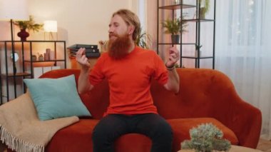 Sakallı, neşeli ve zengin bir adam el hareketini ziyan ediyor ya da etrafa para saçıyor, daha fazla bahşiş, daha fazla kar, piyango, vücut dili, paylaşım. Evde, kanepede oturan genç, kızıl saçlı adam.