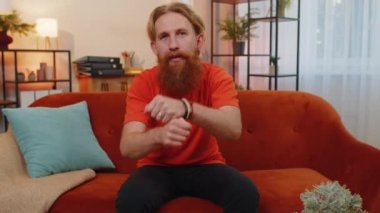 Gibi. Mutlu sakallı genç adam onaylayıcı bir şekilde kameraya bakıyor olumlu bir şey işaret ediyor, iyi haber, olumlu geri bildirim gibi. Yetişkin bir adam evdeki turuncu koltukta oturuyor.