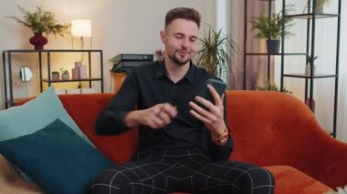 Kanepede oturan yetişkin bir adamın portresi modern ev dairesinde cep telefonu gülümsemesi kullanıyor. Çevrimiçi akıllı telefon sosyal medya uygulamaları üzerinden mesajlaşan genç adam, rahatlama filmi izliyor