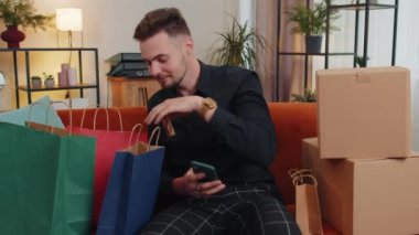 Happy shopaholic consumer man sitting with shopping bags at modern home apartment making online order, payment with credit plastic bank card. Young guy on couch at home. Discount sale on internet