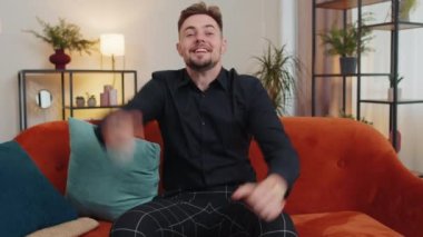 Happy caucasian man shouting, celebrating success, winning, goal achievement, good news, raising fists in gesture I did it, lottery luck at home apartment. Young guy indoors in living room on couch