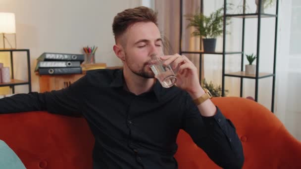 一个口渴可爱的男人坐在室内 手里拿着一杯天然的水 他的肖像让他喝了一口静水 防止脱水 有良好生活习惯 健康体形 体重减轻观念的人 — 图库视频影像